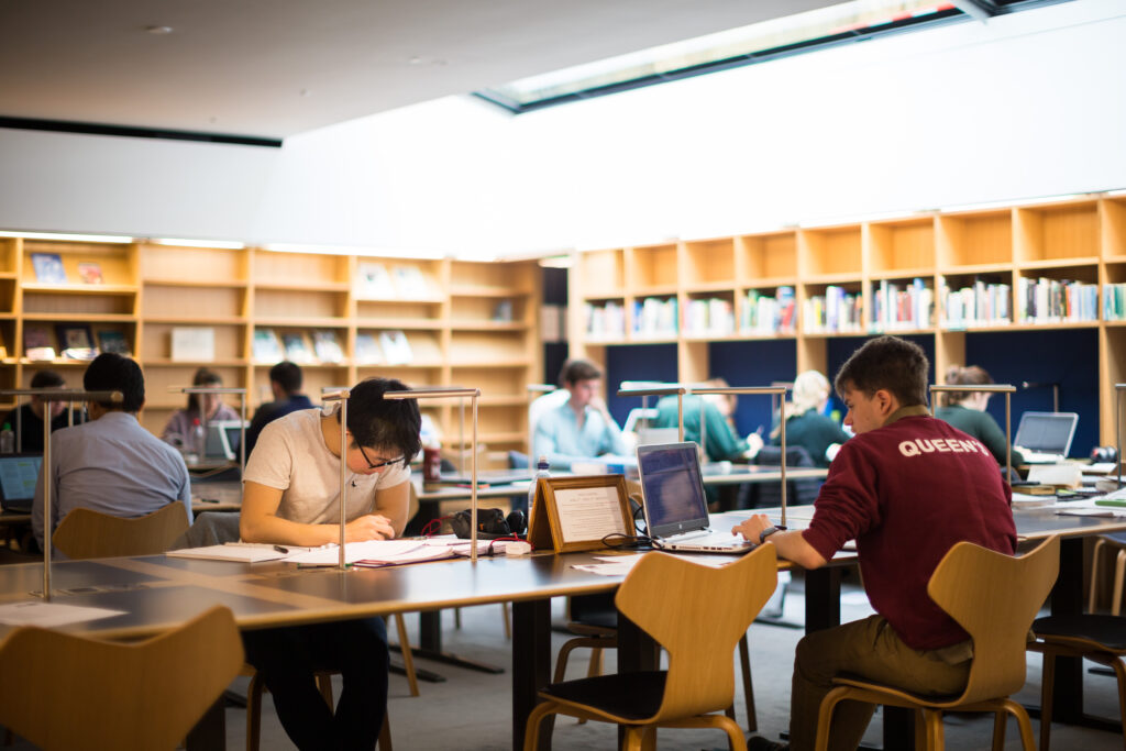 Photograph of the modern New Library reading room area, showing three rows of desks with students sitting at them working. There are pale wooden bookcases in the background, and bright light coming in from a skylight above the right-hand part of the reading room.