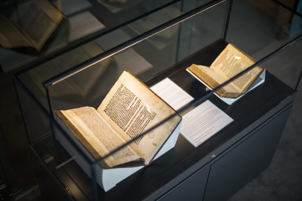 Photograph of a small exhibition case located in the New Library. The case is glass-topped and holds two antique books mounted in cradles so the pages are open. There are accompanying text panels, and the exhibition case lights illuminate the books.