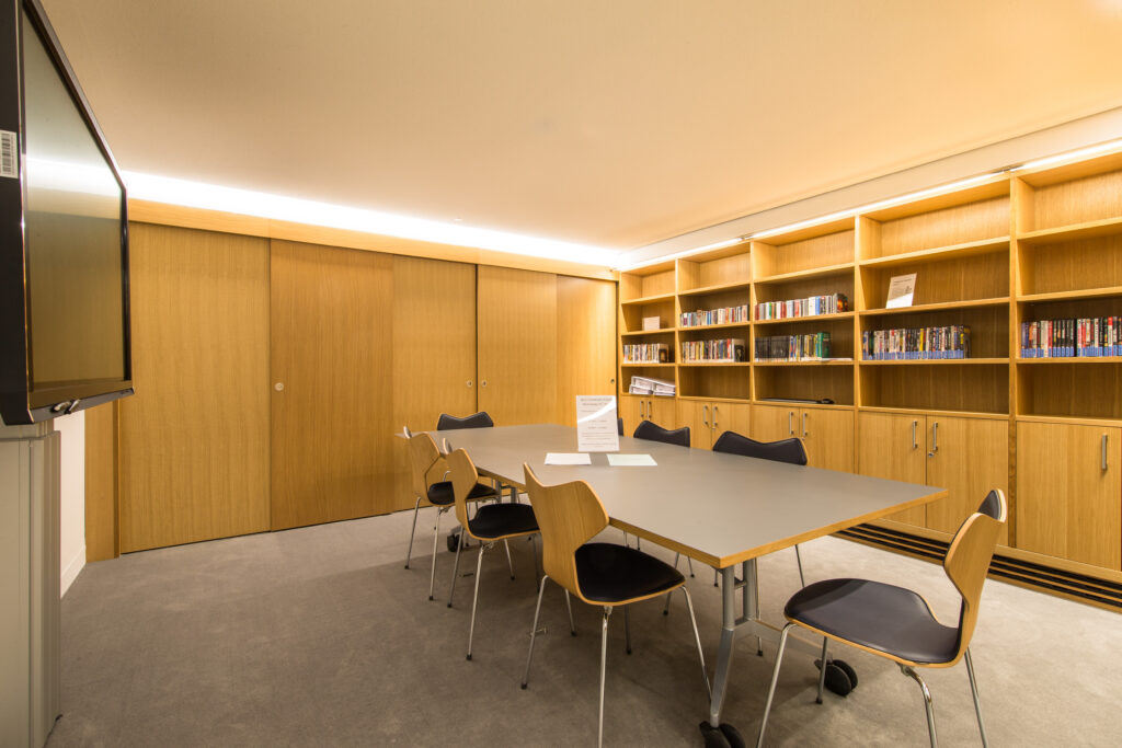 Photograph of the interior of the Multi-Purpose Room. The room has a large central table with six chairs arranged around it. On the wall to the left is a large display screen. On the shelves to the right are DVDs and CDs that can be borrowed.