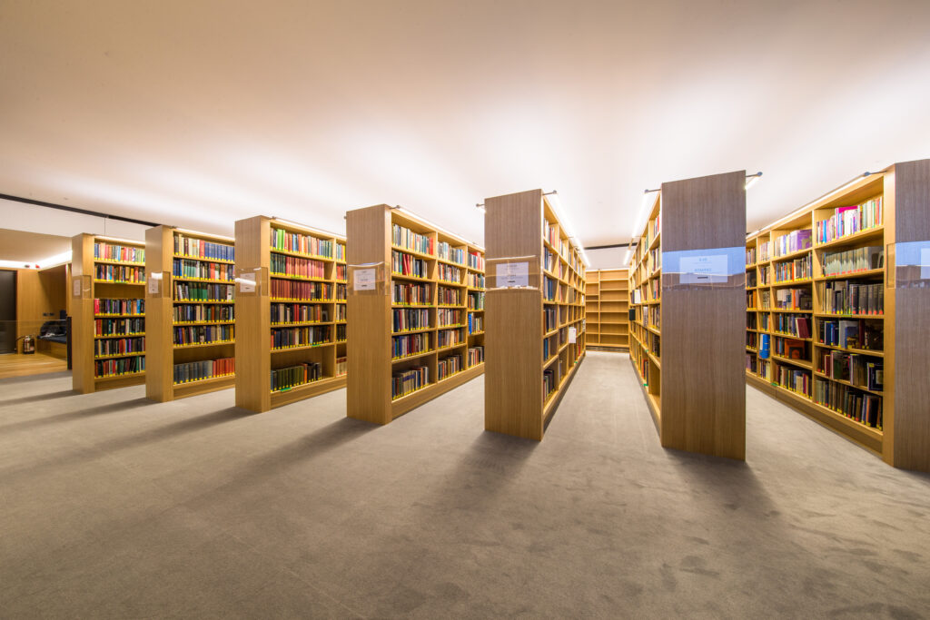 A wide-angle photograph of the book shelving in the New Library, seen from the end of the rows of shelves so they appear to fan out. The shelves are pale wood and filled with rows of colourful modern book spines.