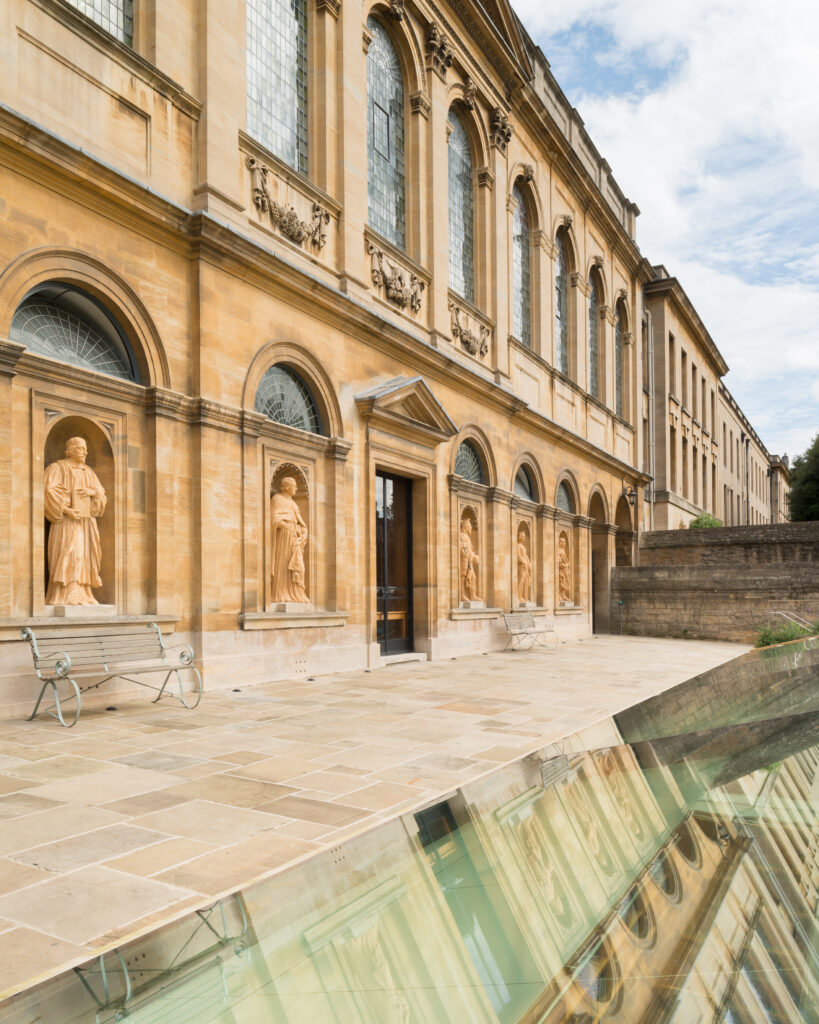 Photograph of the exterior of the Queen’s Library, seen from within the garden of the Provost’s Lodgings. The building is of neo-classical/baroque design. The ground level of this elevation features a series of niches with full-length sculptures of figures important to the College’s history within them. Around the sculptures are arches, supported by pilasters. In the centre of the façade is a door topped by a pediment. The second storey of the building is partially out of frame, but features a similar series of arches, this time framing large windows.