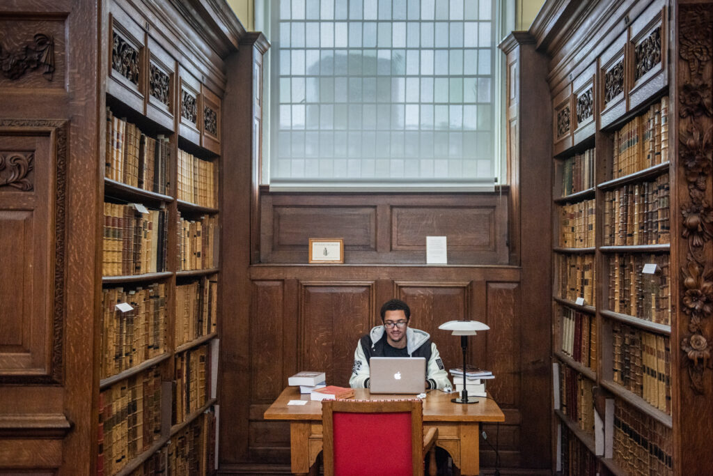 Photograph of a student seated at a library desk, working at a laptop. There are books and papers stacked around him and tall, dark wooden bookcases with rows of leather bound old books on either side of him, and a large window behind him.