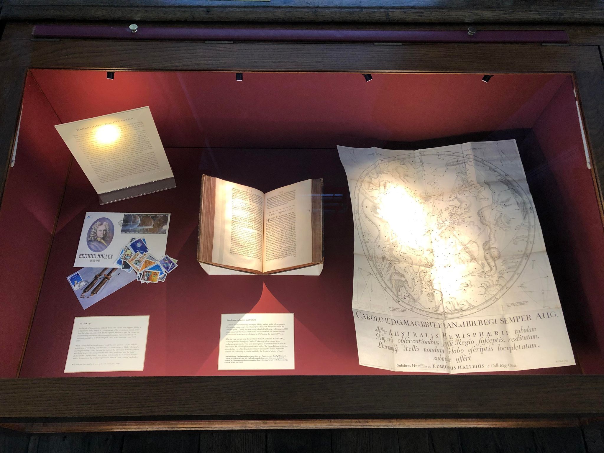 An exhibition case containing ephemera, a mounted book and a large celestial map, plus captions, against a red fabric background.