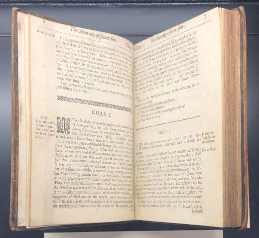 The book 'The anatomy of secret sins...' mounted in a cradle so its pages are open. The pages bare paragraphs of type in a Roman face, with decorative dividers marking new sections. There is one illustrated initial visible on the left-hand page: a woodcut 'I' surrounded by swirling decoration.