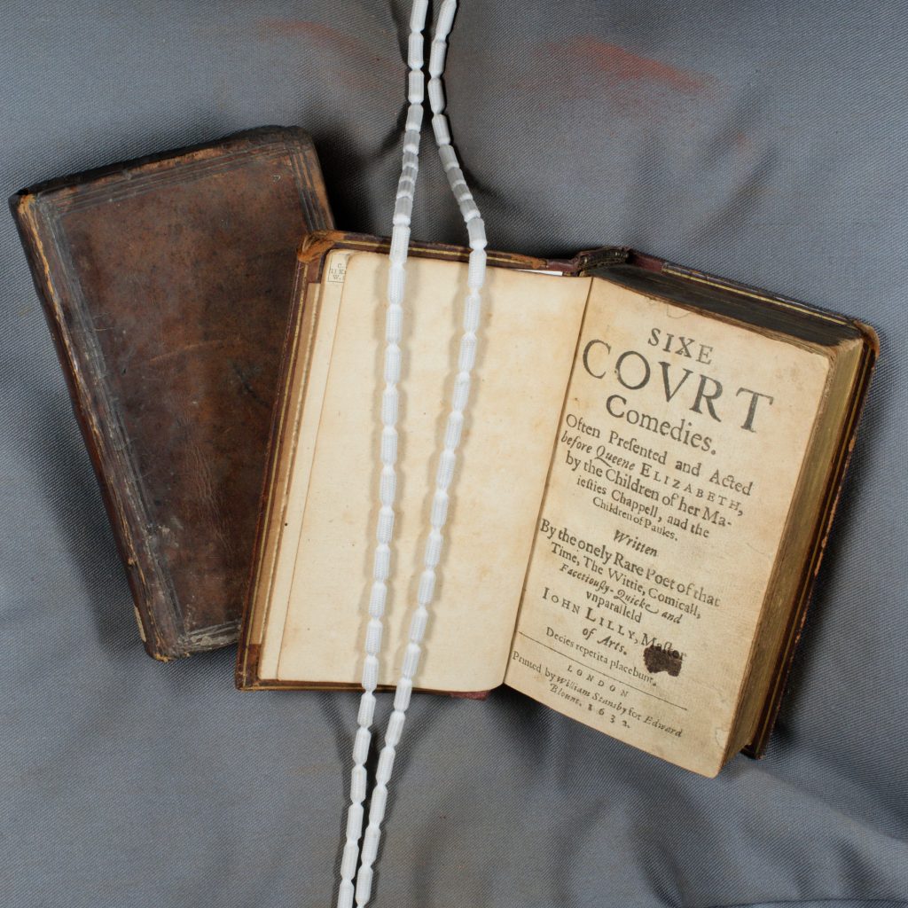 Image shows Sixe Court Comedies, a 1632 book that collects six comedies written by Lyly performed in front of Queen Elizabeth. The book lies open to the title page, on a book support cushion, and is held in place by two weighted 'book snakes' draped across one page.