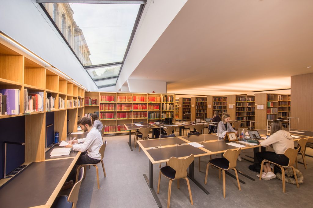 A photograph of the modern New Library reading room with students working at some of the desks. A row of desks runs along the wall to the left, underneath the skylight, and other rows of desks are in the centre of the reading room. Bookcases run around the outside of the space, with further bookcases visible in the background. They are full of colourful modern books.