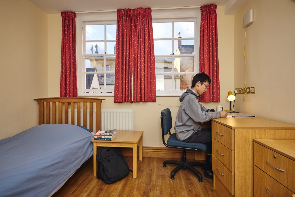 Carrodus Quad bedroom student bedroom with bed, small table, desk, drawers and two windows. A student sits at his desk writing. Through the window you can see the Queen's College main building