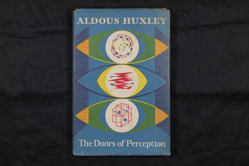 The book 'The Doors of Perception' lying flat so the cover is face-up. The cover has a blue background, with three large abstract eye designs taking up most of the space. They have geometric designs within the pupils and have red, yellow, green, and orange details so each eye is different. The author and title are at the head and foot of the cover respectively, printed in white.