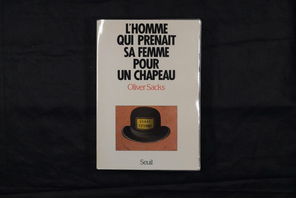 The book 'L’Homme qui prenait sa femme pour un chapeau' lying flat so the cover is faceup. The cover is white, with the author and title printed in a large, bold black font. It bears a small illustration of a black bowler hat against an orange background. The cover is inside a plastic library dust-jacket and so has a slight sheen.