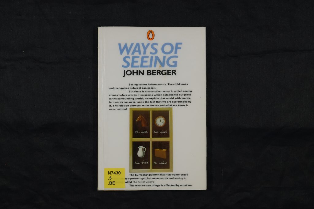A copy of the book 'Ways of Seeing' lying flat so the cover is face-up. The title is in large, bold blue italic font against a white background. An excerpt from the book is printed in small black font towards the centre of the cover. There is a small illustration of a horse, a clock, a milk jug, and a briefcase underneath.