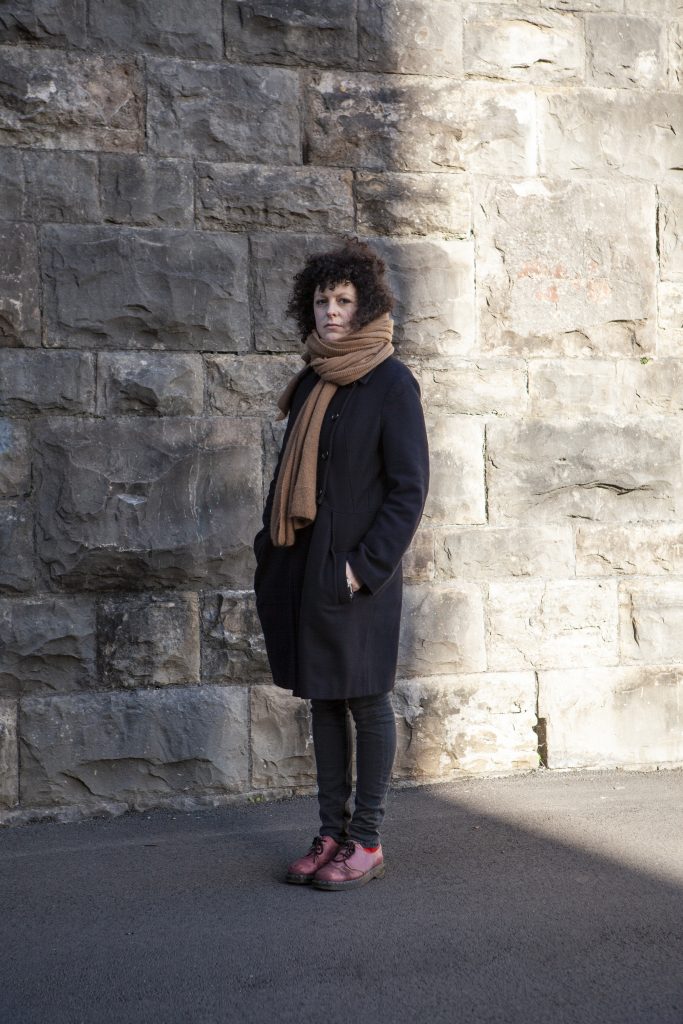 Polly Barton profile photo: Polly is standing in front of a tall stone wall. She is wearing a smart black coat, black trousers, a brown scarf and brown shoes. Her hands are in her pockets and she is looking at the camera.